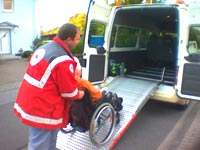 Mitarbeiter des DRK schiebt Rollstuhlfahrerin in das Auto