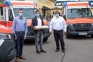 Drei Männer mit Maske vor drei Rettungswagen, ein Mann hält ein großes Blech Kuchen