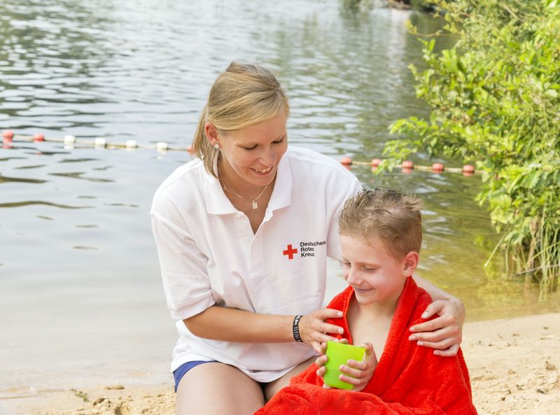 Sanitäterin legt an einem Badesee einem Jungen ein rotes Handtuch um.