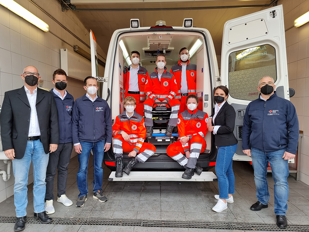 Bild größer ansehen Die fünf neuen Azubis - sitzend im geöffneten Rettungswagen - mit dem Team Ausbildung Rettungsdienst