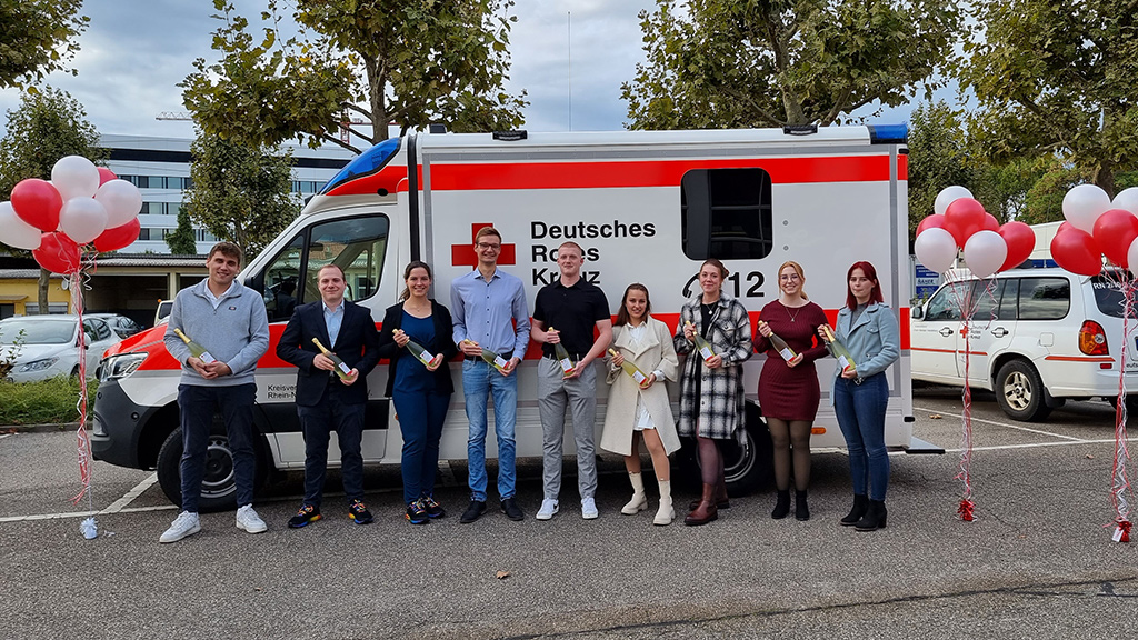 Bild größer ansehen Gruppenbild: 9 Absolventen vor einem Rettungswagen