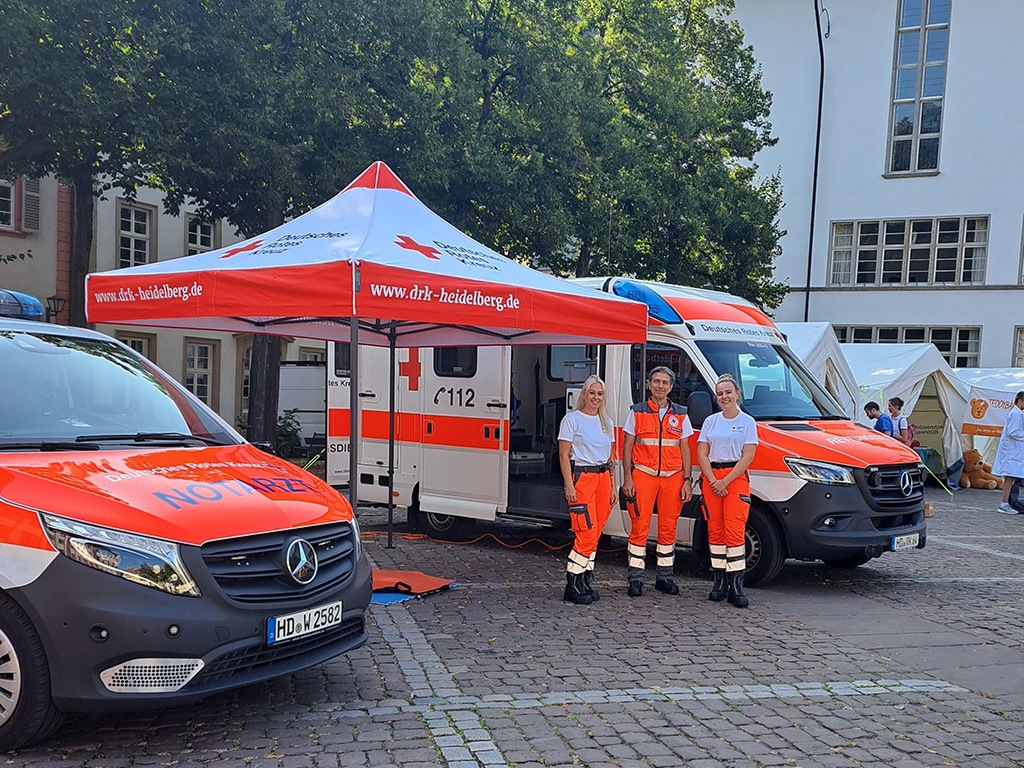 Zwei Sanitäterinnen und ein Sanitäter neben DRK-Rettungsfahrzeugen und einem aufgebauten DRK-Sonnenschirm