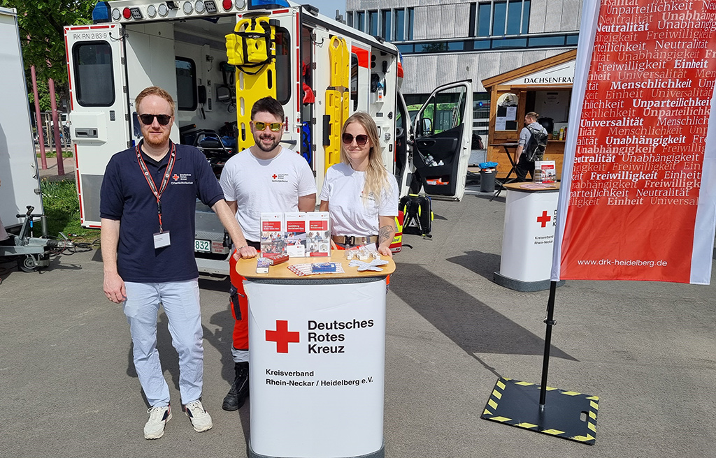 Felix Zurbrüggen mit Jonas Jänsch und Michelle Pfeifer an einem DRK-Stand vor einem geöffneten Rettungswagen