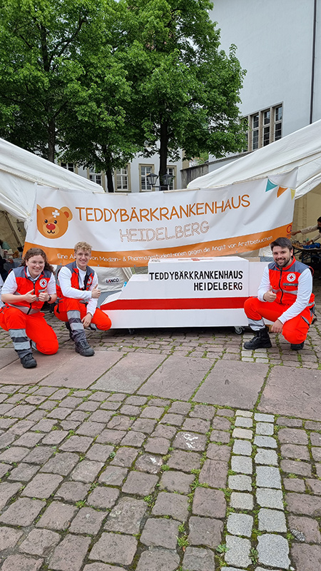 Bild größer ansehen Drei DRK Notfallsanitäter/innen vor einem Banner mit der Aufschrift Teddybärenkrankenhaus Heidelberg