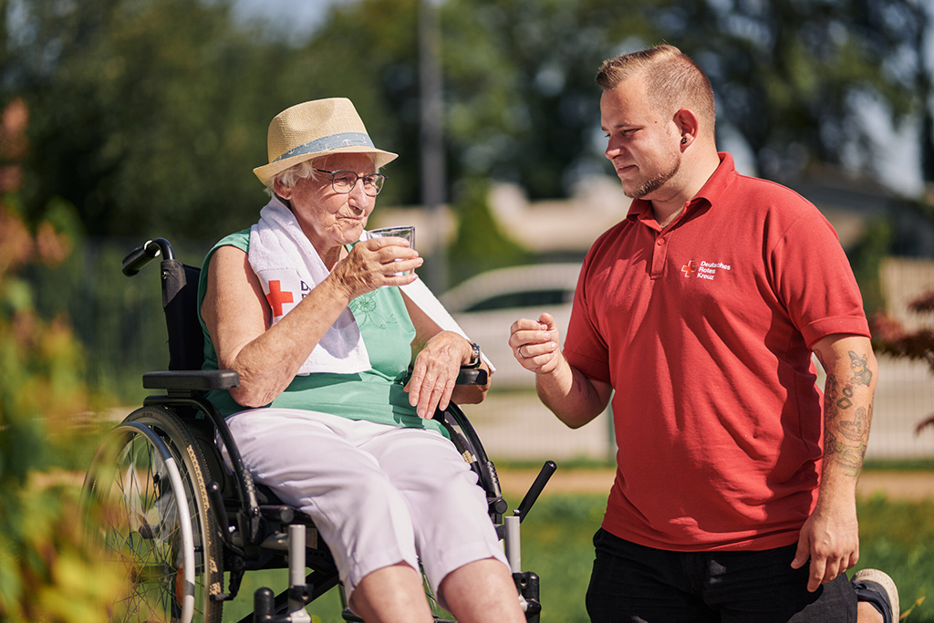Bild größer ansehen Überhitzte Seniorin im Rollstuhl erhält von einem DRK-Mitarbeiter ein Glas Wasser