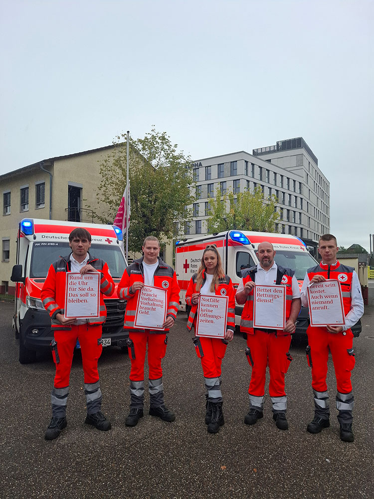 Team der Rettungswache Heidelberg vor zwei Rettungswagen halten Zettel hoch, auf denen sie aufmerksam machen, dass die Notfallreform nachgebessert werden soll.