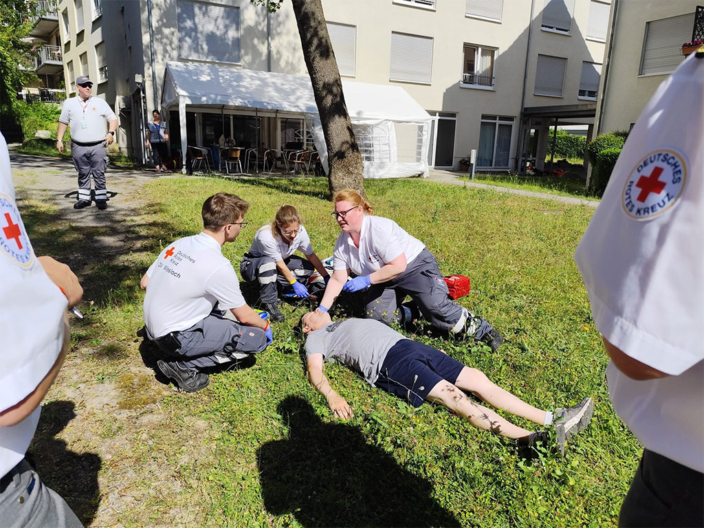 Erste Hilfe-Übung: DRK-Helfer kümmern sich um einen kollabierten jungen Mann, der auf dem Rasen liegt