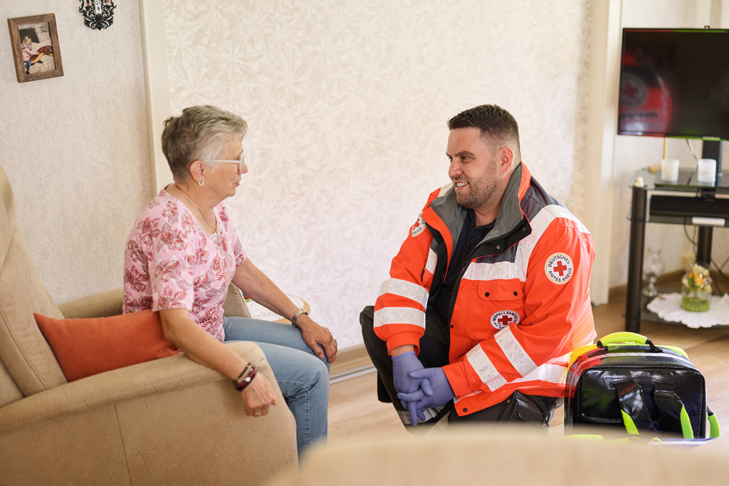 Sanitäter hockt neben einer älteren Dame, die auf einem Sessel in ihrer Wohnung sitzt